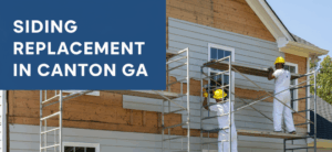 Three construction workers in yellow hard hats and white overalls are on scaffolding, installing light grey siding on a house. Part of the house still shows exposed wooden sheathing. A blue tarp on the green lawn is covered with siding materials and tools.